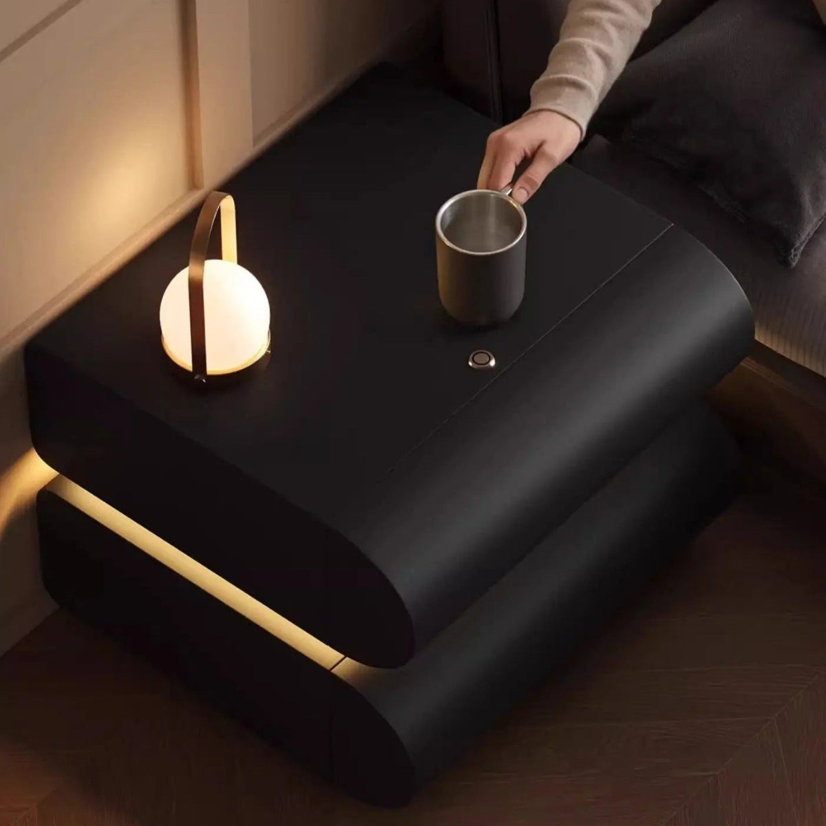 Aura Smart LED luxury nightstand in black with warm LED strip glow between tiers shown from overhead with a model reaching for a mug and portable lamp on top — LivDecor
