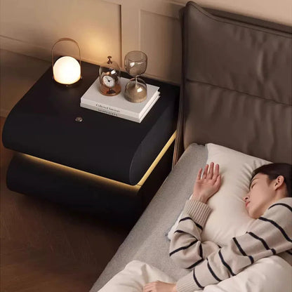 Aura Smart LED luxury nightstand in black with warm LED strip glow shown from overhead beside a sleeping model in a cozy luxury bedroom styled with a portable lamp, clock, and hourglass — LivDecor
