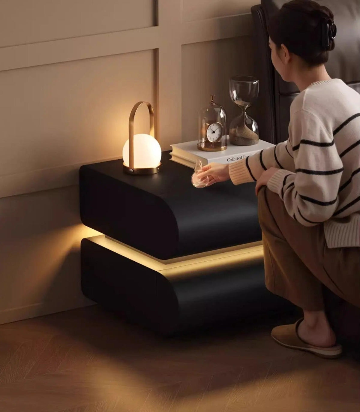 Aura Smart LED luxury nightstand in black tech leather with warm LED base glow styled in a moody bedroom with a model reaching for a glass beside a portable lamp and hourglass — LivDecor
