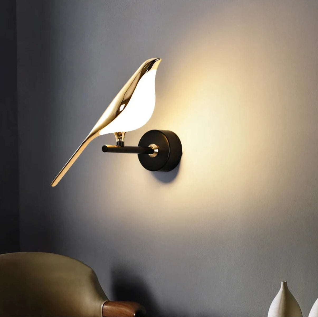 Avis Modern Origami Bird LED wall sconce in gold and matte black single-bird configuration illuminated close-up showing warm halo glow on grey wall with olive green lounge chair and white ceramic vases — LivDecor
