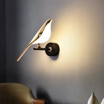 Avis Modern Origami Bird LED wall sconce in gold and matte black single-bird configuration illuminated close-up showing warm halo glow on grey wall with olive green lounge chair and white ceramic vases — LivDecor
