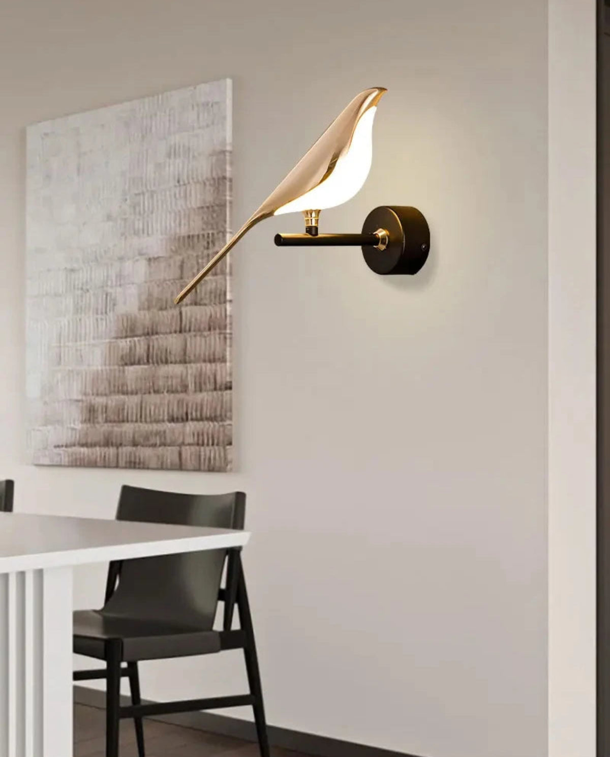 Avis Modern Origami Bird LED wall sconce in gold and matte black single-bird configuration illuminated on a white wall beside a textured abstract canvas above a white dining table with black folding chairs — LivDecor
