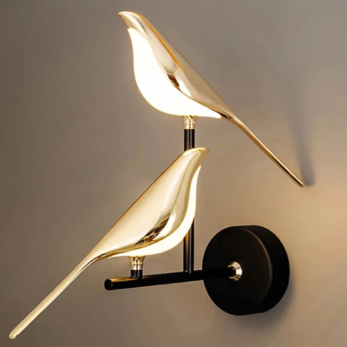 Avis Modern Origami Bird LED wall sconce in gold and matte black two-bird configuration illuminated close-up showing both birds glowing with warm light on staggered arms against a warm taupe wall — LivDecor
