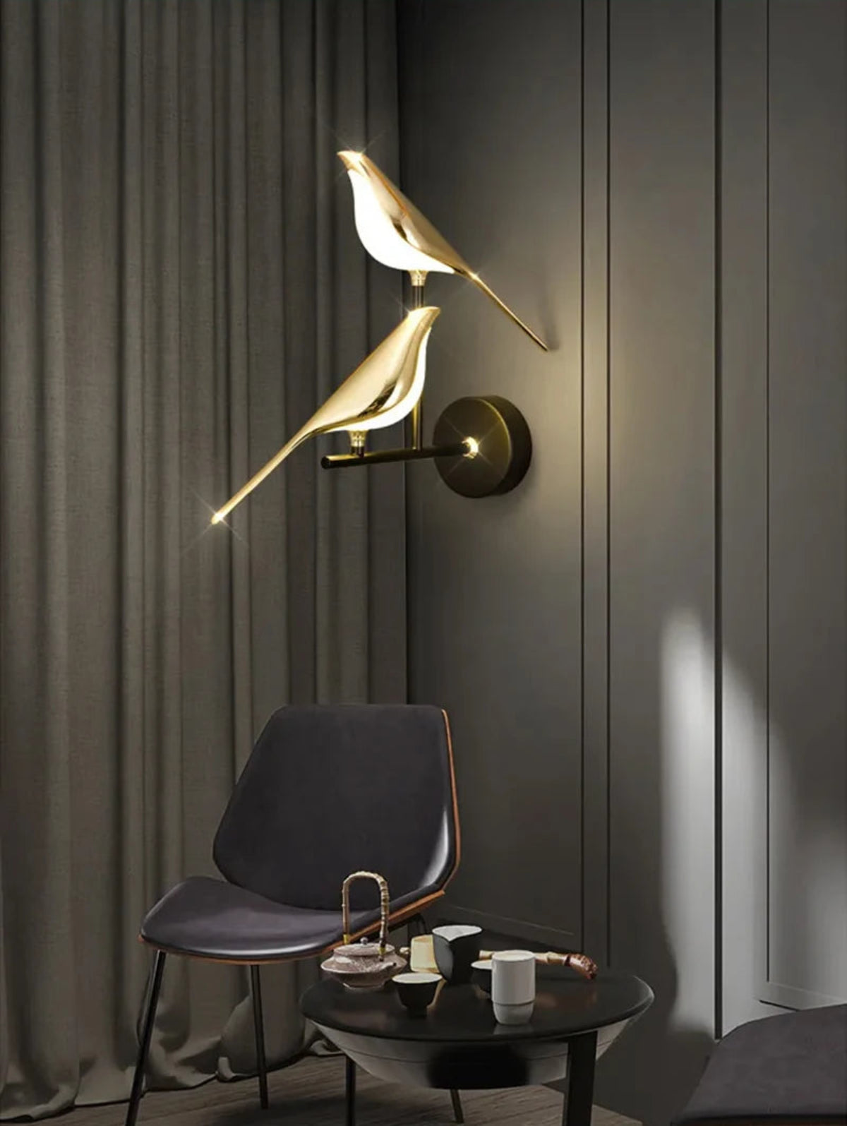 Avis Modern Origami Bird LED wall sconce in gold and matte black two-bird configuration illuminated on a dark grey fluted panel wall with floor-length drapes, slate blue lounge chair, and black round side table with tea set — LivDecor
