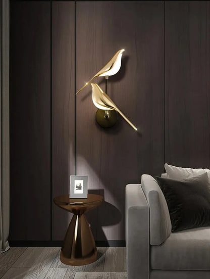 Avis Modern Origami Bird LED wall sconce in gold and matte black two-bird configuration illuminated against a dark walnut wood panel wall with sculptural walnut side table and grey sofa — LivDecor

