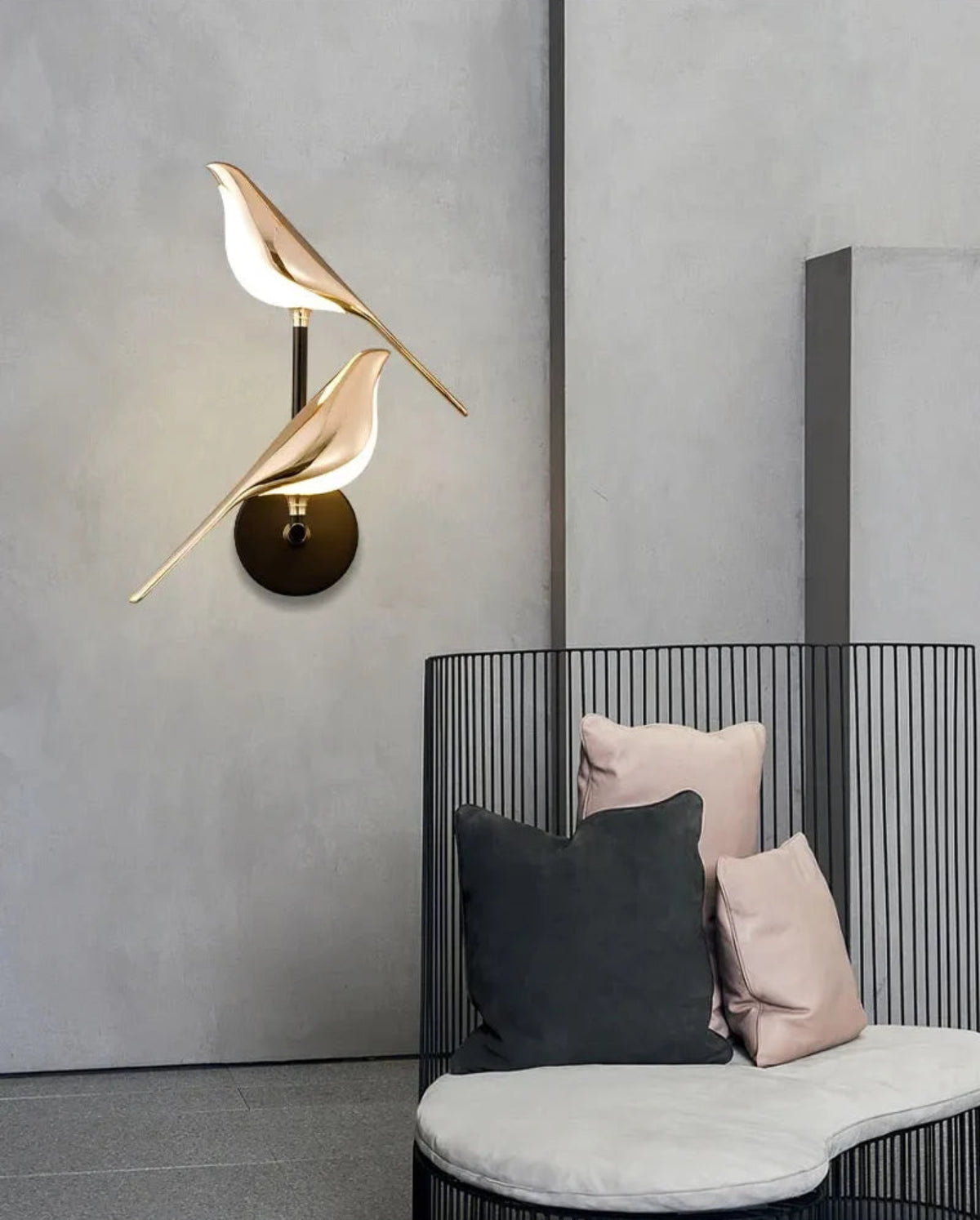 Avis Modern Origami Bird LED wall sconce in gold and matte black two-bird configuration illuminated on a raw concrete wall beside a curved black wire lounge chair with blush and charcoal cushions — LivDecor
