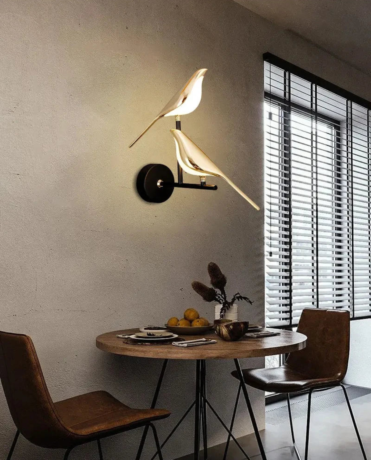 Avis Modern Origami Bird LED wall sconce in gold and matte black two-bird configuration illuminated on a raw plaster wall above a round walnut bistro table with tan leather chairs and slatted blinds — LivDecor
