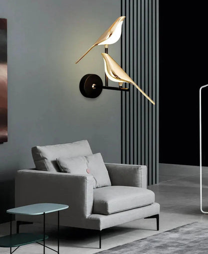 Avis Modern Origami Bird LED wall sconce in gold and matte black two-bird configuration illuminated on a slate grey fluted panel wall above a light grey lounge chair with teal side table — LivDecor
