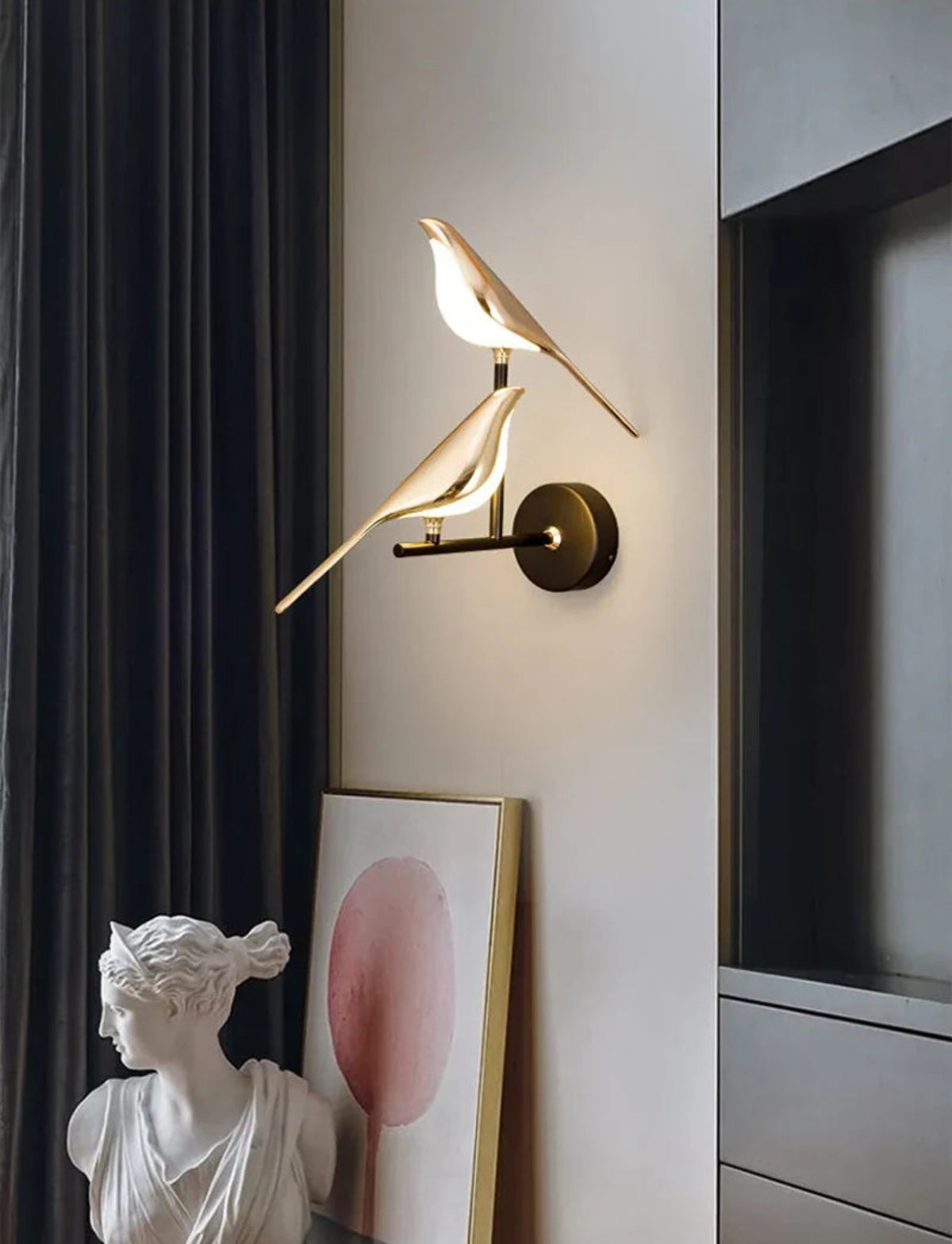 Avis Modern Origami Bird LED wall sconce in gold and matte black two-bird configuration illuminated on a warm white wall with charcoal drapes, classical Venus bust, and gold-framed abstract print — LivDecor
