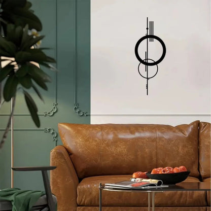 Black metal geometric abstract wall art with double circle and vertical line design mounted on a cream wall above a tan leather sofa in an eclectic living room with sage green panelling and a tropical plant — LivDecor