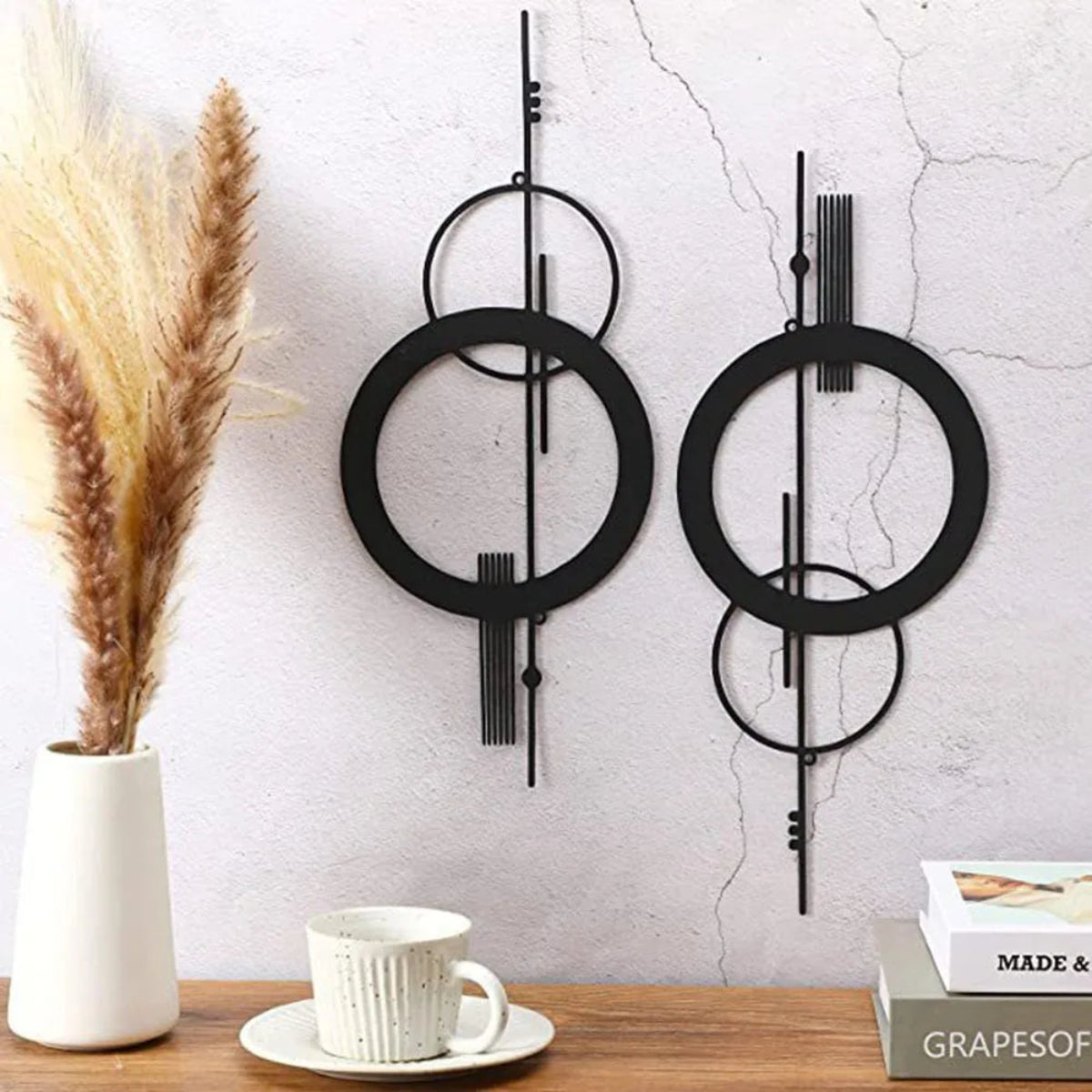 Two black metal geometric abstract wall art pieces with double circle and vertical line design displayed as a pair on a textured plaster wall, styled with dried pampas grass in a white vase and a coffee cup on a wooden shelf — LivDecor
