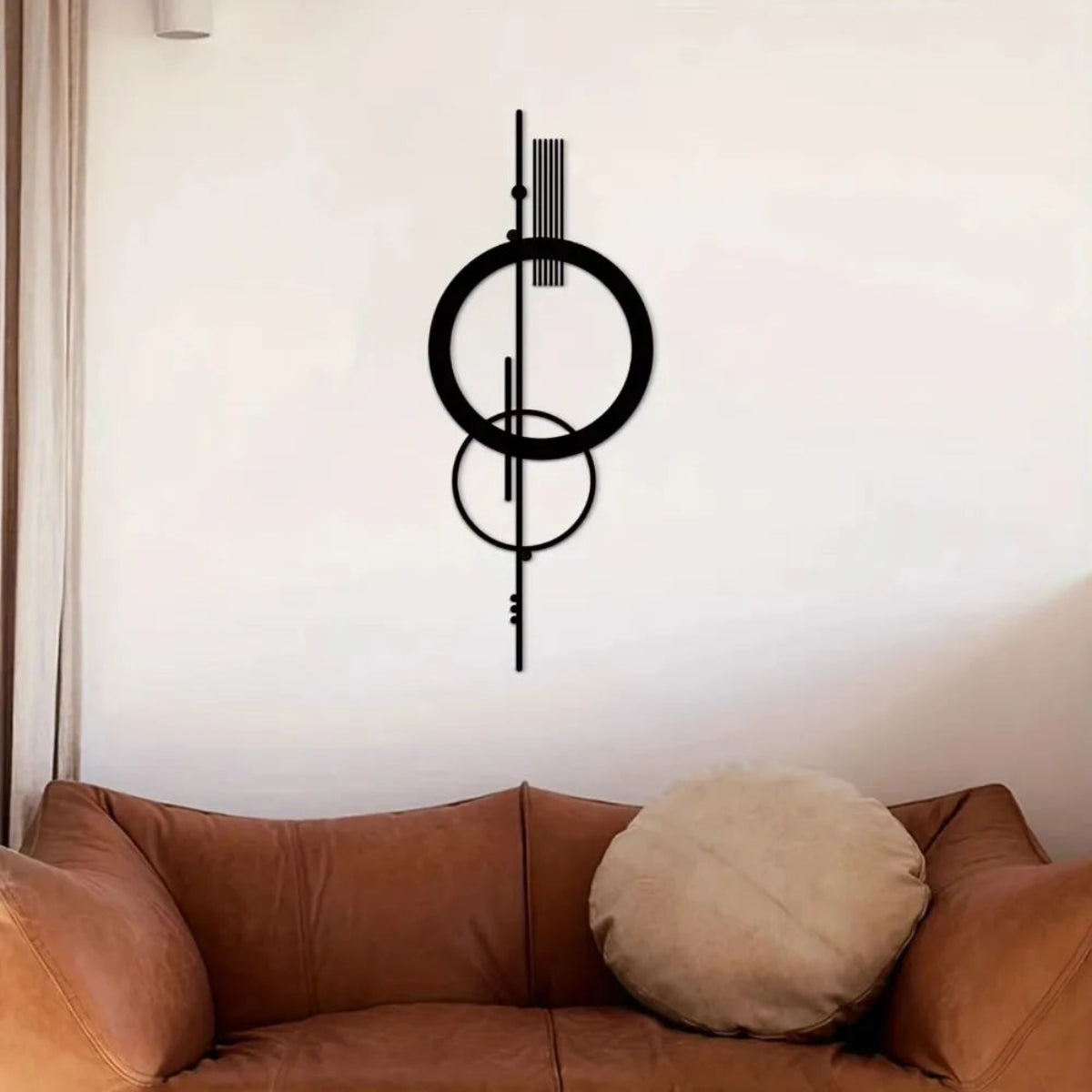 Black metal geometric abstract wall art with double circle and vertical line design mounted on a white wall above a tan leather sofa with a round beige cushion — LivDecor
