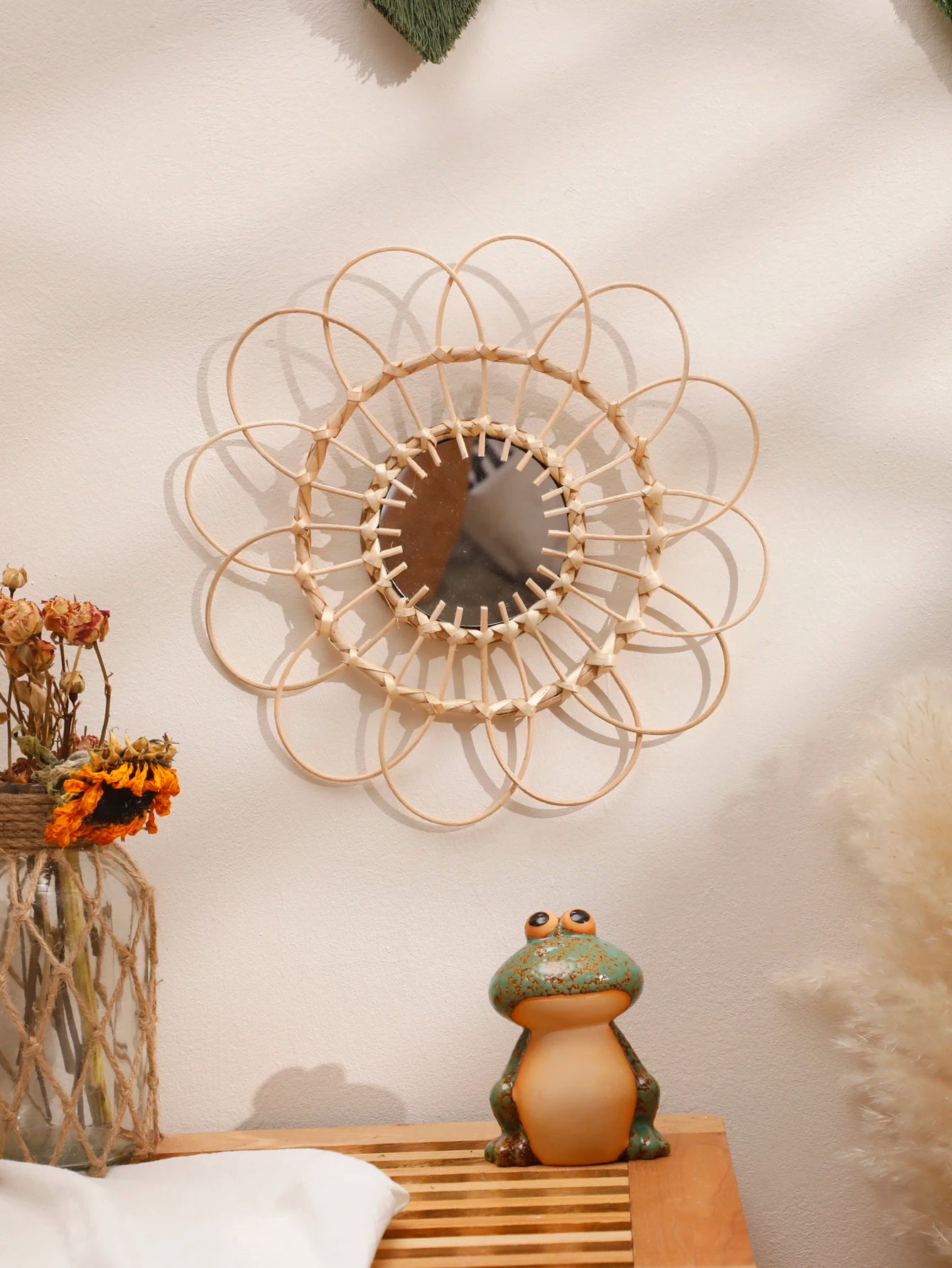 Boho rattan sunflower wall mirror on a cream wall above a wooden shelf with a glass vase of dried roses and sunflowers and a ceramic frog figurine, with pampas grass to the right — LivDecor
