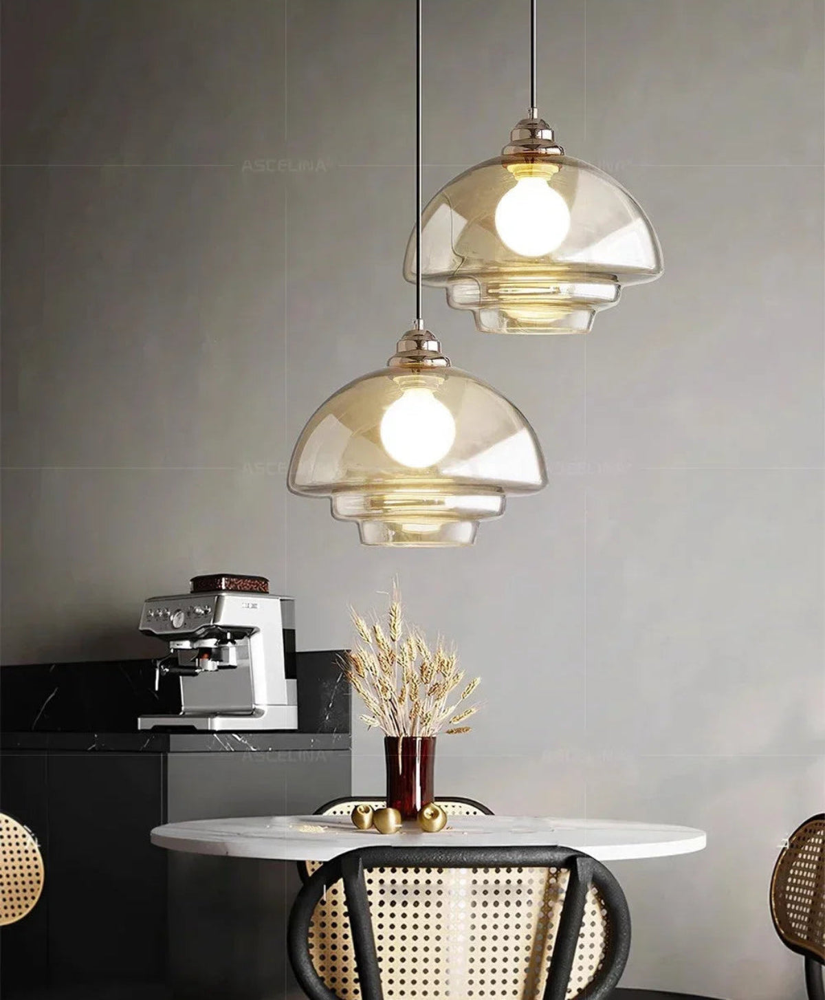 Two clear amber tiered glass pendant lights with chrome fittings hung above a round marble dining table with rattan chairs, dried wheat stems in a vase, and an espresso machine in the background — LivDecor