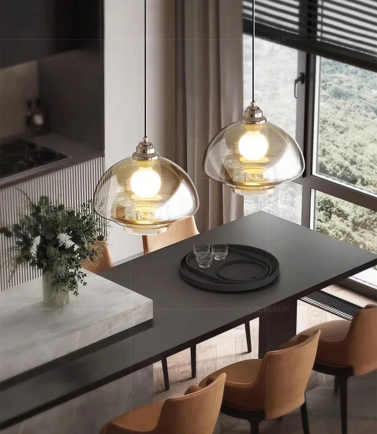 Two clear amber tiered glass pendant lights with chrome fittings illuminated above a dark dining table with caramel leather chairs, a marble island with eucalyptus bouquet, and a forest view through floor-to-ceiling windows — LivDecor