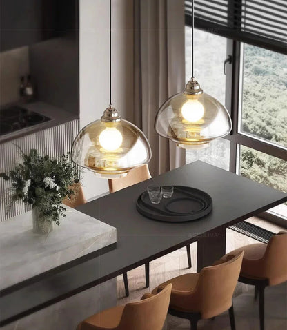 Two clear amber tiered glass pendant lights with chrome fittings illuminated above a dark dining table with caramel leather chairs, a marble island with eucalyptus bouquet, and a forest view through floor-to-ceiling windows — LivDecor