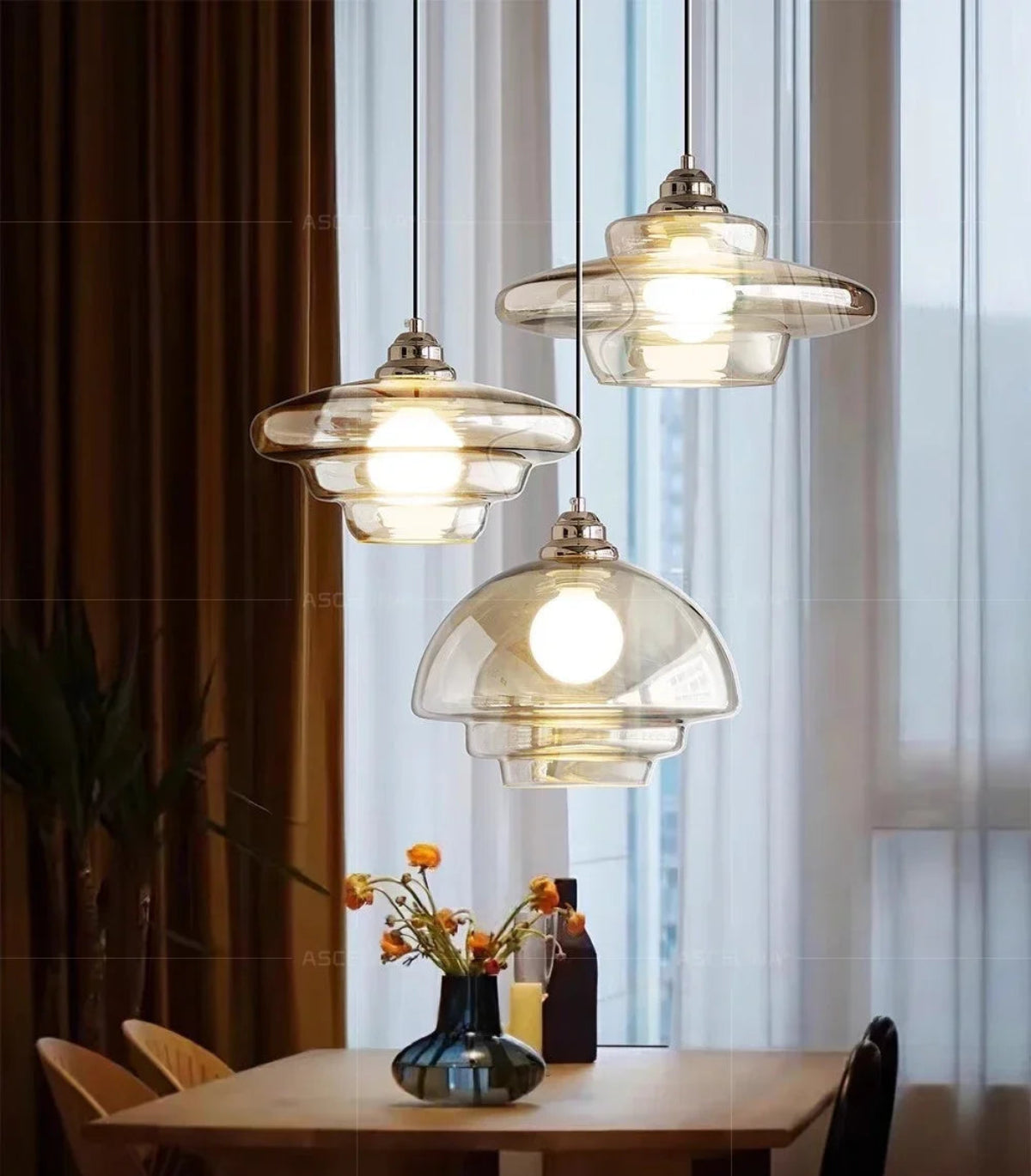 Three clear amber tiered glass pendant lights in saucer, dome, and flat tiered shapes hung above a wood dining table with orange ranunculus flowers in a blue vase and sheer curtains — LivDecor