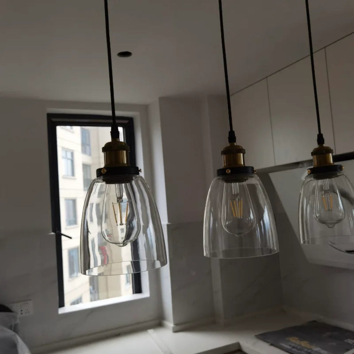 Three clear glass bell pendant lights with brass fittings installed above a kitchen counter in natural daylight with an urban window view — LivDecor