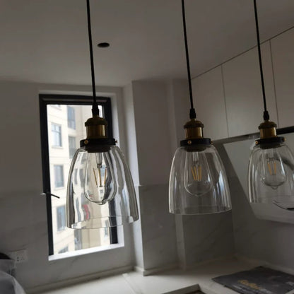 Three clear glass bell pendant lights with brass fittings installed above a kitchen counter in natural daylight with an urban window view — LivDecor