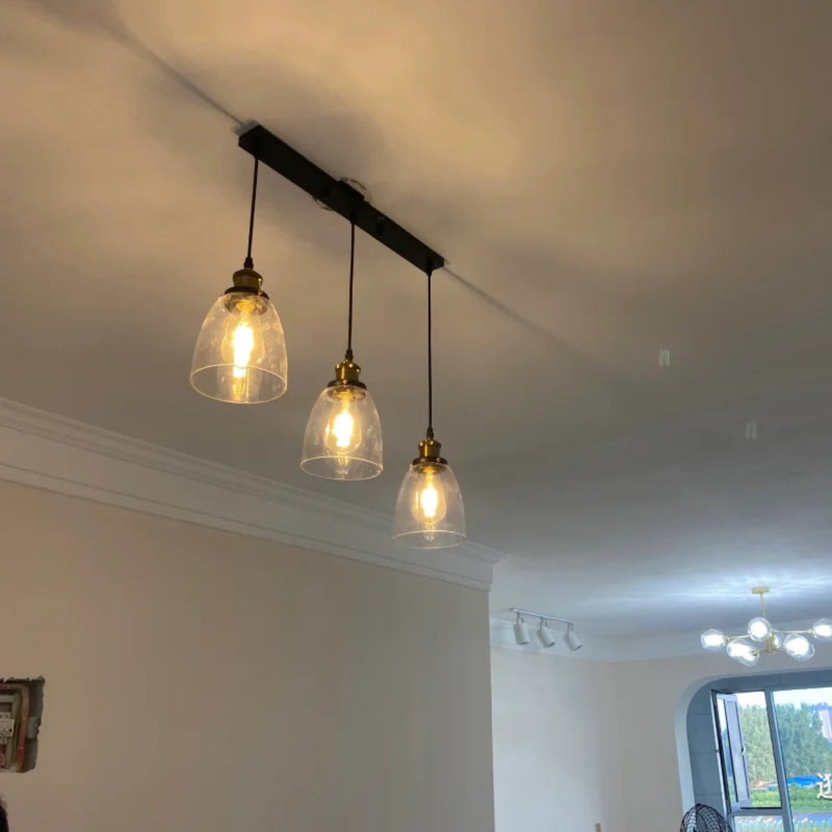 Three clear glass bell pendant lights with brass fittings hung at staggered heights from a black long bar ceiling plate in a real home installation — LivDecor