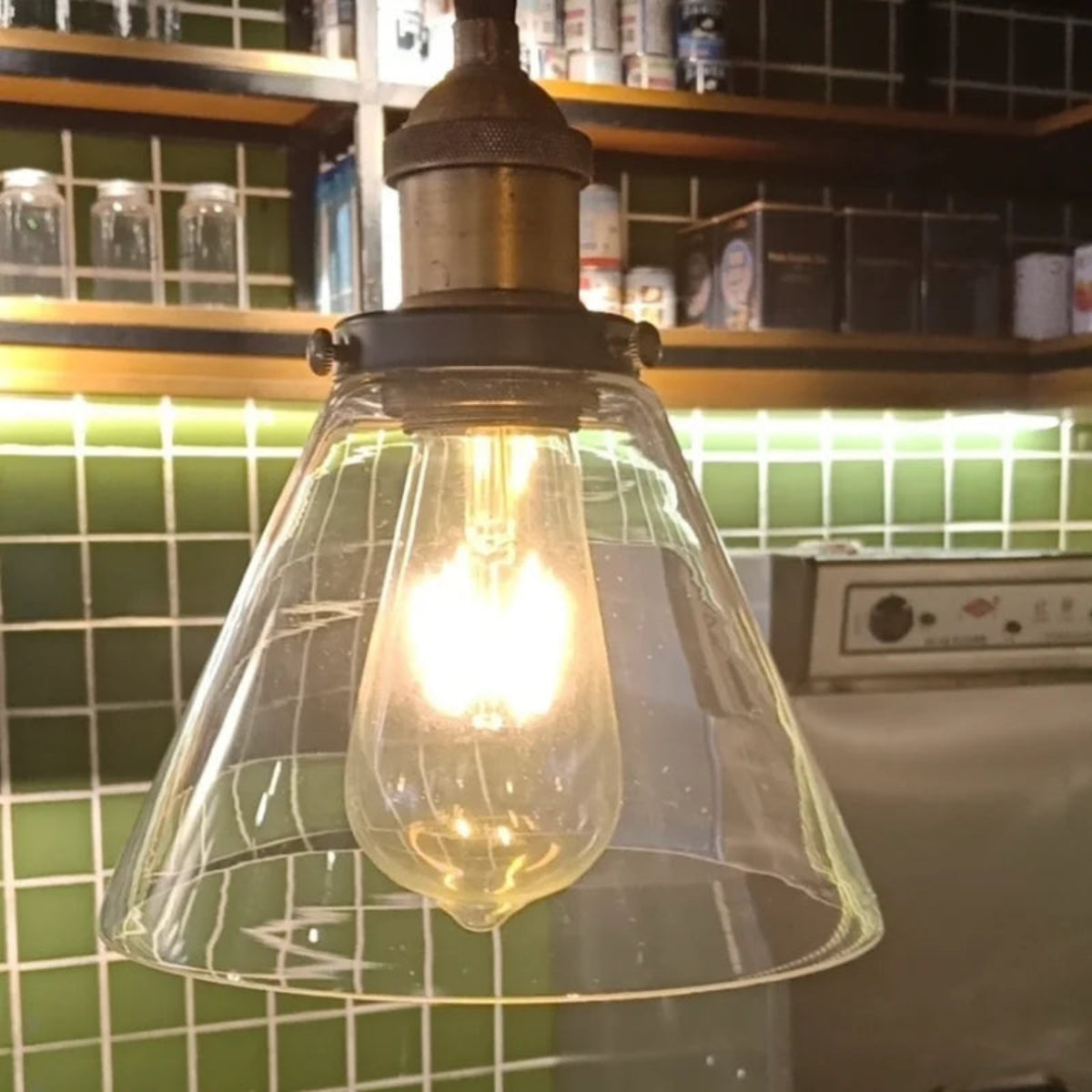Clear glass cone pendant light with brass fitting glowing with an Edison filament bulb in a bar or café setting with green subway tiles and wooden shelving in the background — LivDecor