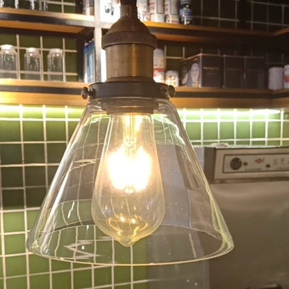 Clear glass cone pendant light with brass fitting glowing with an Edison filament bulb in a bar or café setting with green subway tiles and wooden shelving in the background — LivDecor