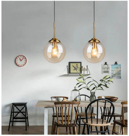 Two clear glass globe pendant lights with gold hardware hanging above a rustic wood dining table with mixed chairs in a bright casual dining room — LivDecor
