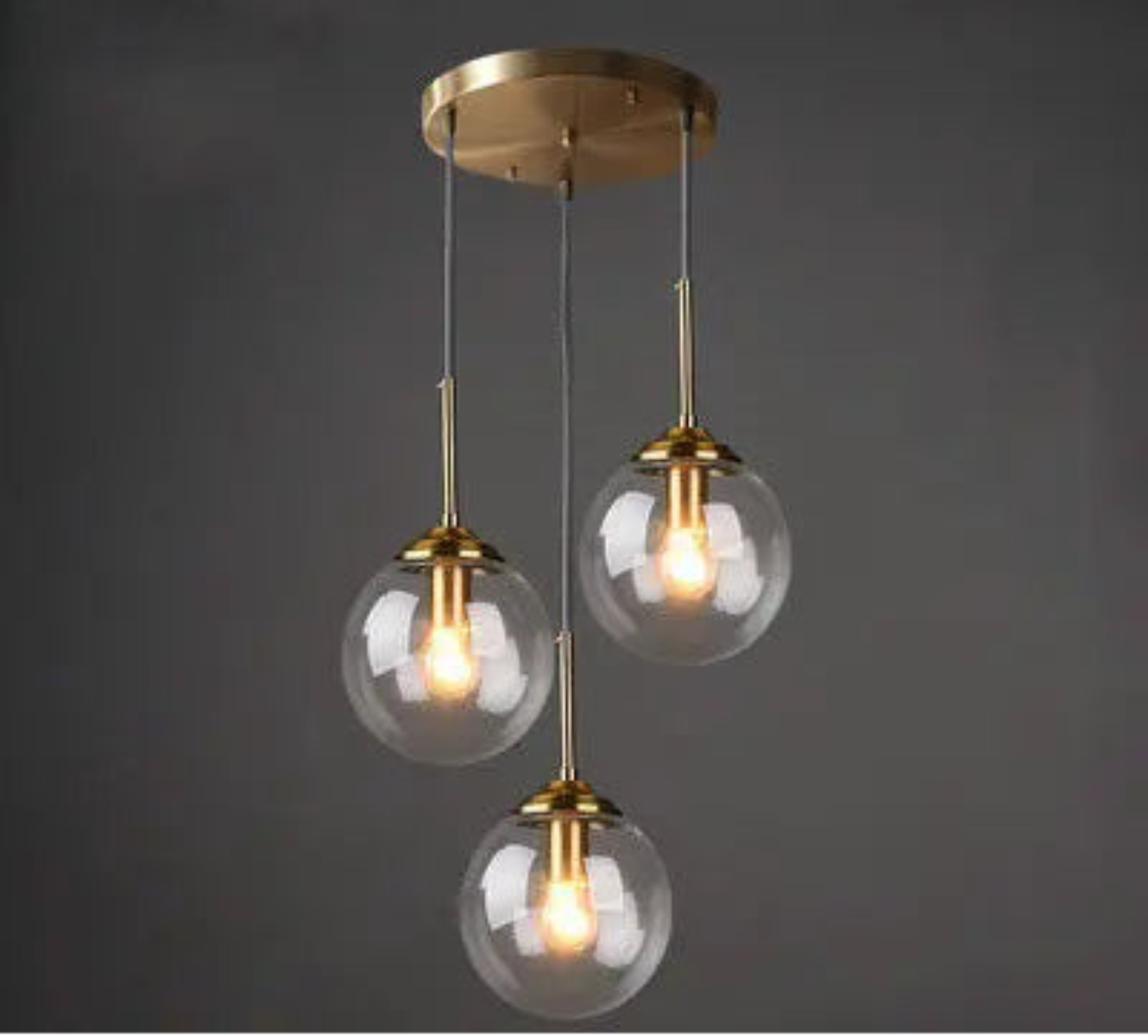 Three clear glass globe pendant lights on staggered gold cords and round ceiling mount in a cluster configuration against a dark grey background — LivDecor