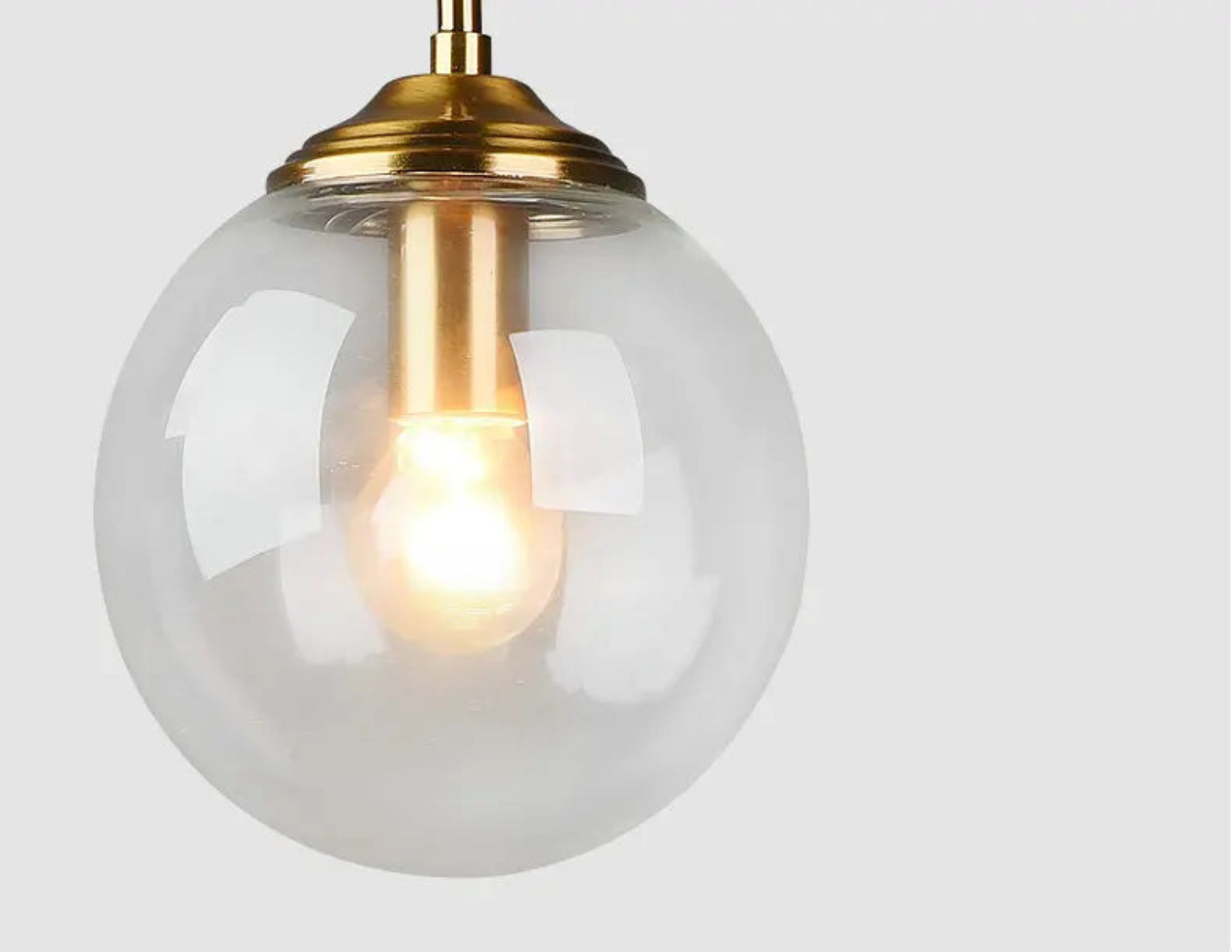Close-up of a clear glass globe pendant light with warm glowing filament bulb and brushed gold hardware fitting against a white background — LivDecor
