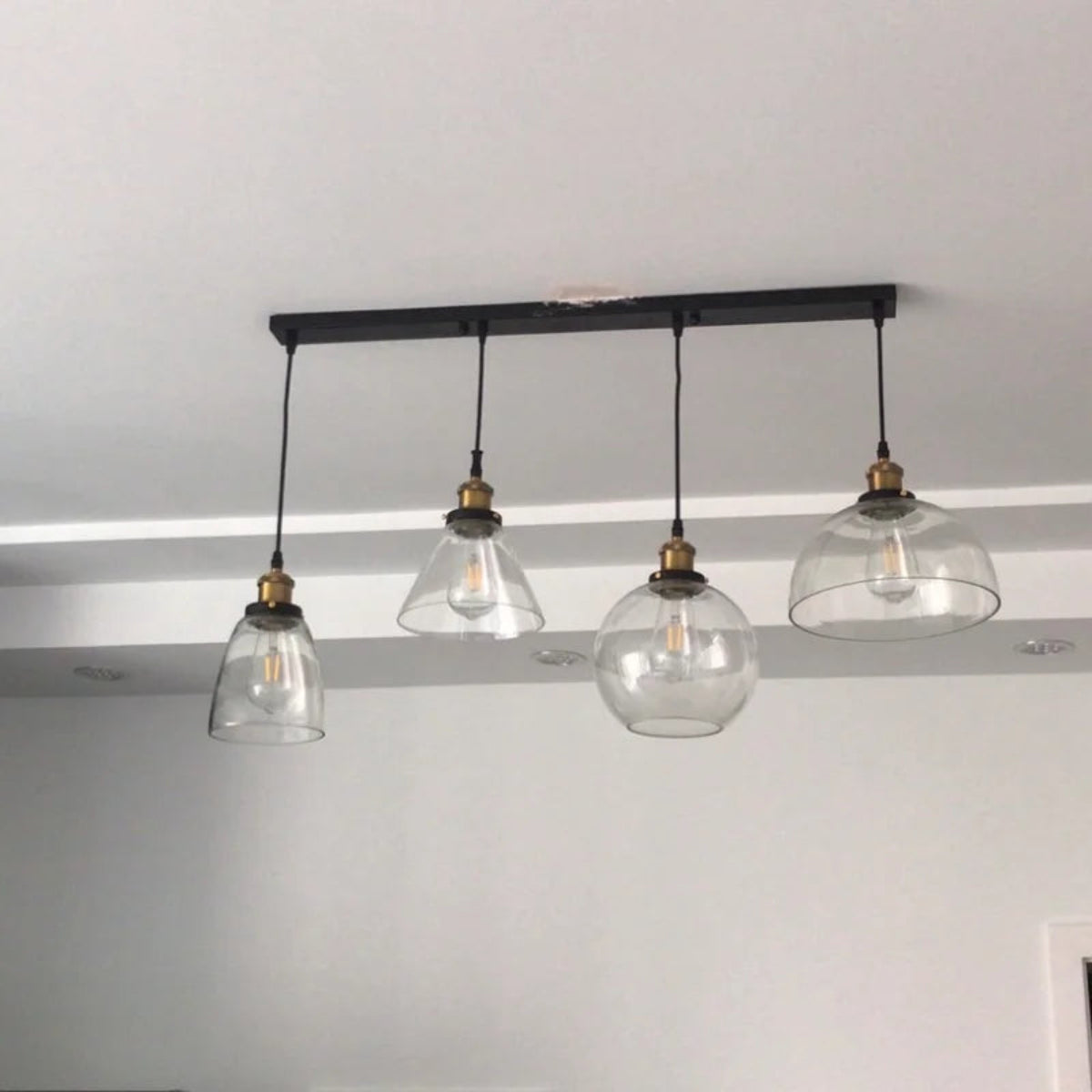 Four clear glass pendant lights in bell, cone, globe and dome shapes hung from a black linear bar ceiling plate in a real home installation — LivDecor