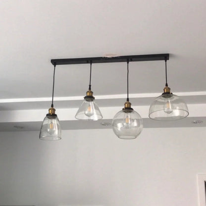 Four clear glass pendant lights in bell, cone, globe and dome shapes hung from a black linear bar ceiling plate in a real home installation — LivDecor