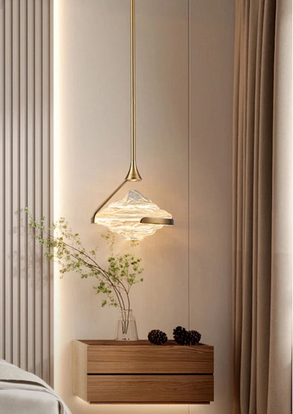 Single clear wave glass geometric pendant light on a gold stem glowing warmly above a wood nightstand with botanical vase beside a fluted wall panel in a luxury bedroom — LivDecor