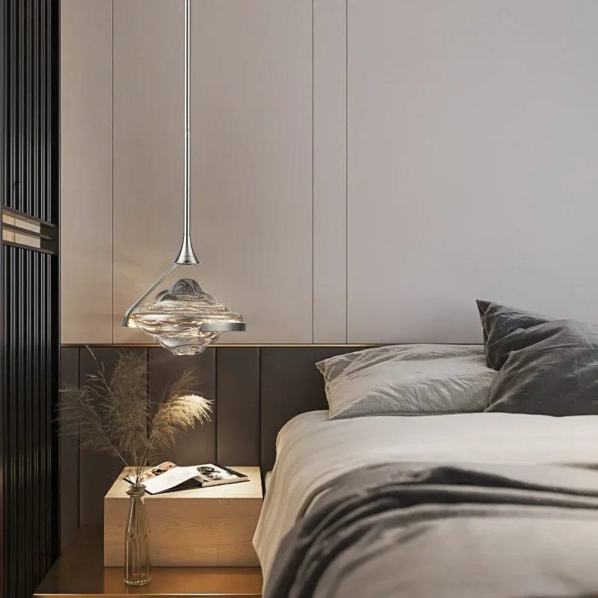 Single clear wave glass geometric pendant light on a chrome stem glowing above a bedside table with pampas grass vase beside a dark wood headboard and grey paneled wall — LivDecor
