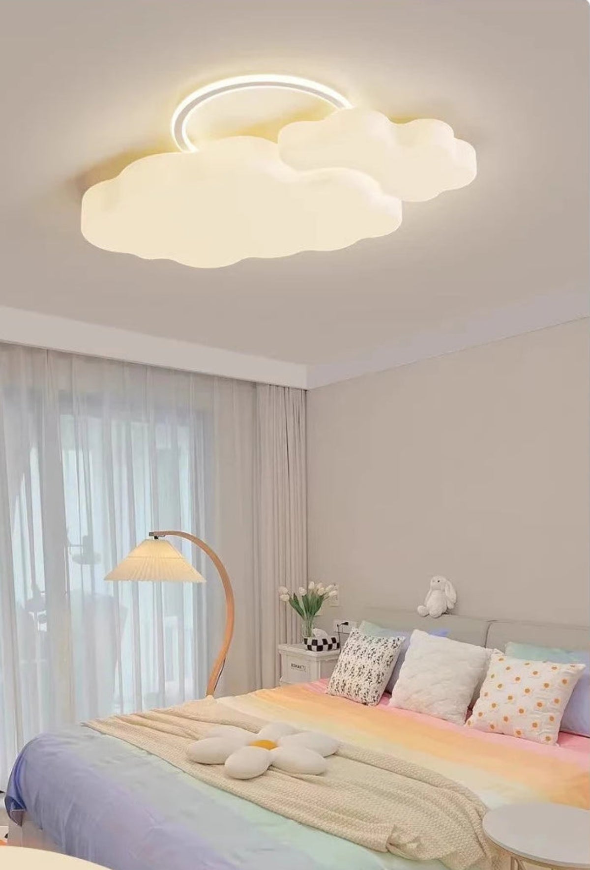 Cumulus Luxe cream sculptural cloud LED ceiling light with halo ring glowing soft white in a kids bedroom with pastel rainbow bedding, tulips, cloud cushions, and wooden arc floor lamp — LivDecor
