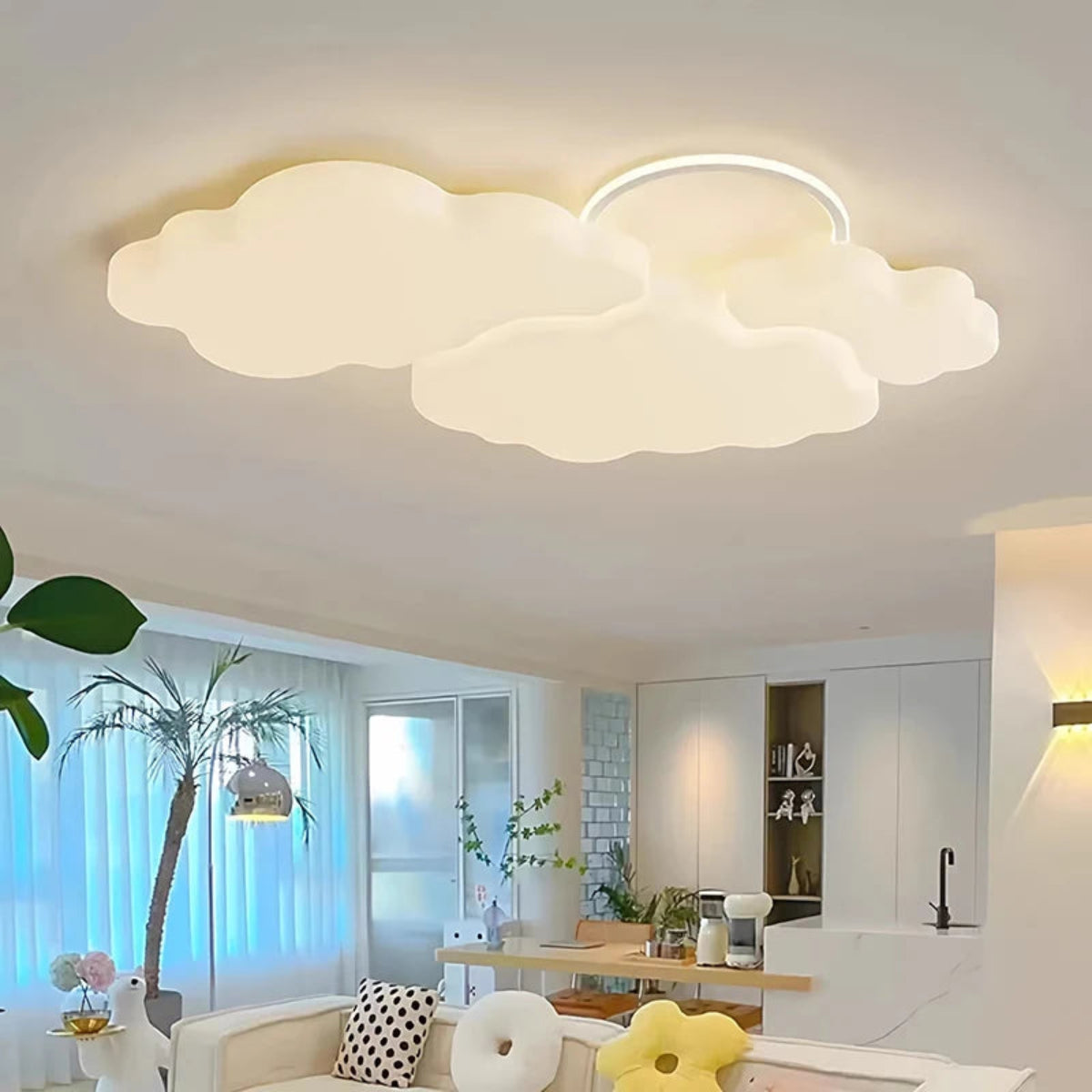 Cumulus Luxe cream sculptural cloud LED ceiling light with halo ring glowing warm white in a bright open-plan living and dining space with palm tree, modern kitchen, and daisy cushions — LivDecor
