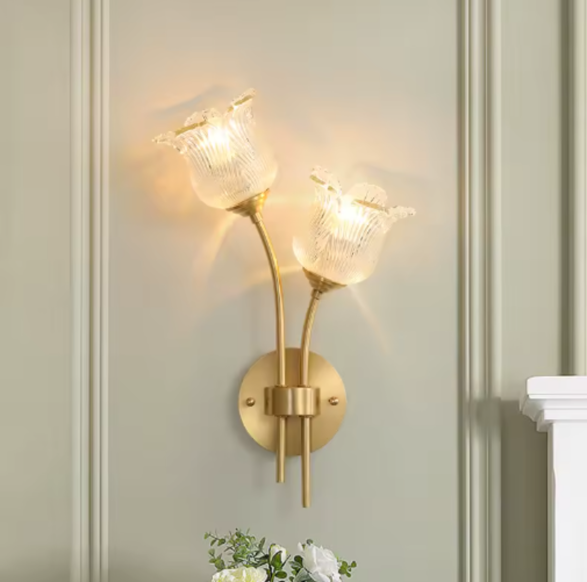 Flora Luxe botanical brass double-head wall sconce with clear ribbed floral glass shades illuminated on a sage-green paneled wall with greenery below, angled right — LivDecor
