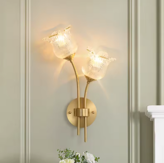 Flora Luxe botanical brass double-head wall sconce with clear ribbed floral glass shades illuminated on a sage-green paneled wall with greenery below, angled right — LivDecor
