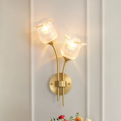 Flora Luxe botanical brass double-head wall sconce with two ruffled floral glass shades illuminated on an ivory paneled wall with roses below, angled right — LivDecor
