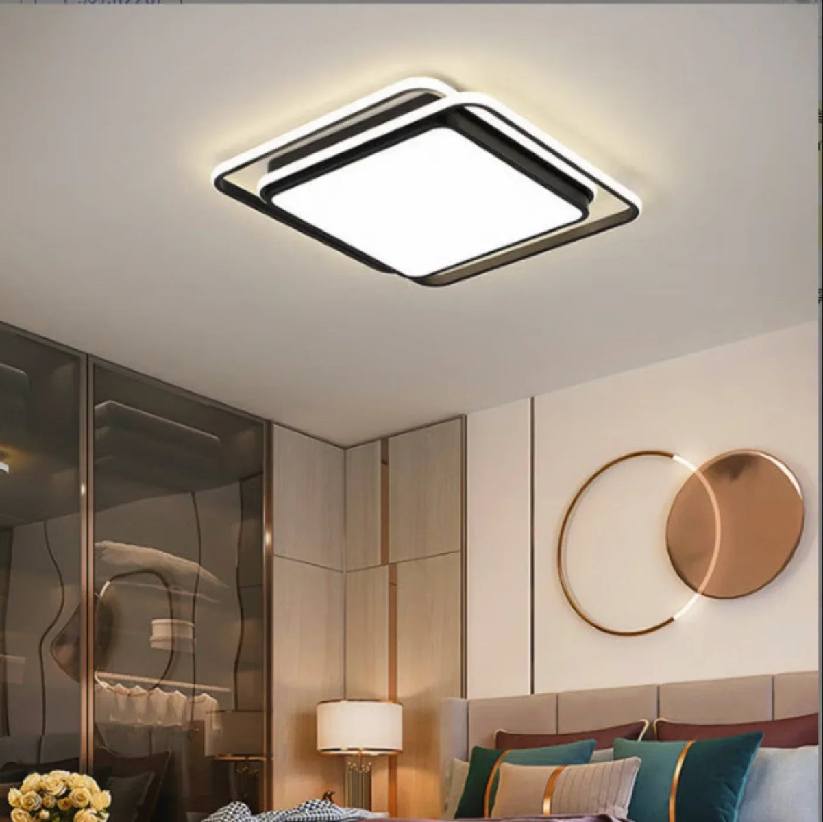 Halo modern tricolor LED flush mount ceiling light in matte black square frame glowing warm white in a gold-accented bedroom with circular wall art, teal cushions, and glass wardrobe — LivDecor
