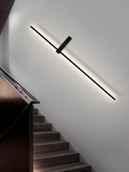 Minimalist linear LED wall sconce in matte black mounted diagonally along a white staircase wall casting a warm halo glow above dark wood steps and a black metal handrail lifestyle shot — LivDecor
