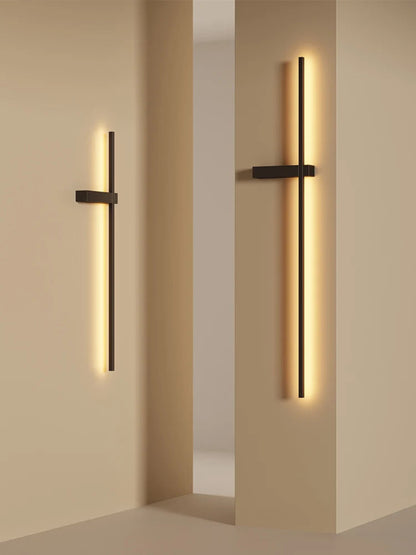 Pair of minimalist linear LED wall sconces in matte black mounted on warm sand-toned architectural columns casting rich amber glow in a modern interior lifestyle shot — LivDecor
