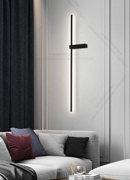 Single minimalist linear LED wall sconce in matte black mounted vertically on a white panelled wall beside a grey sofa with navy herringbone drapes in a modern living room lifestyle shot — LivDecor

