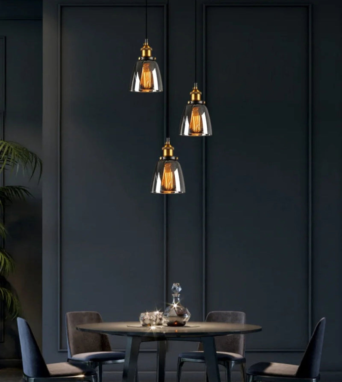 Three smoke grey glass bell pendant lights with brass fittings hung at staggered heights above a dark round dining table with velvet chairs and a crystal decanter against dark teal paneled walls — LivDecor