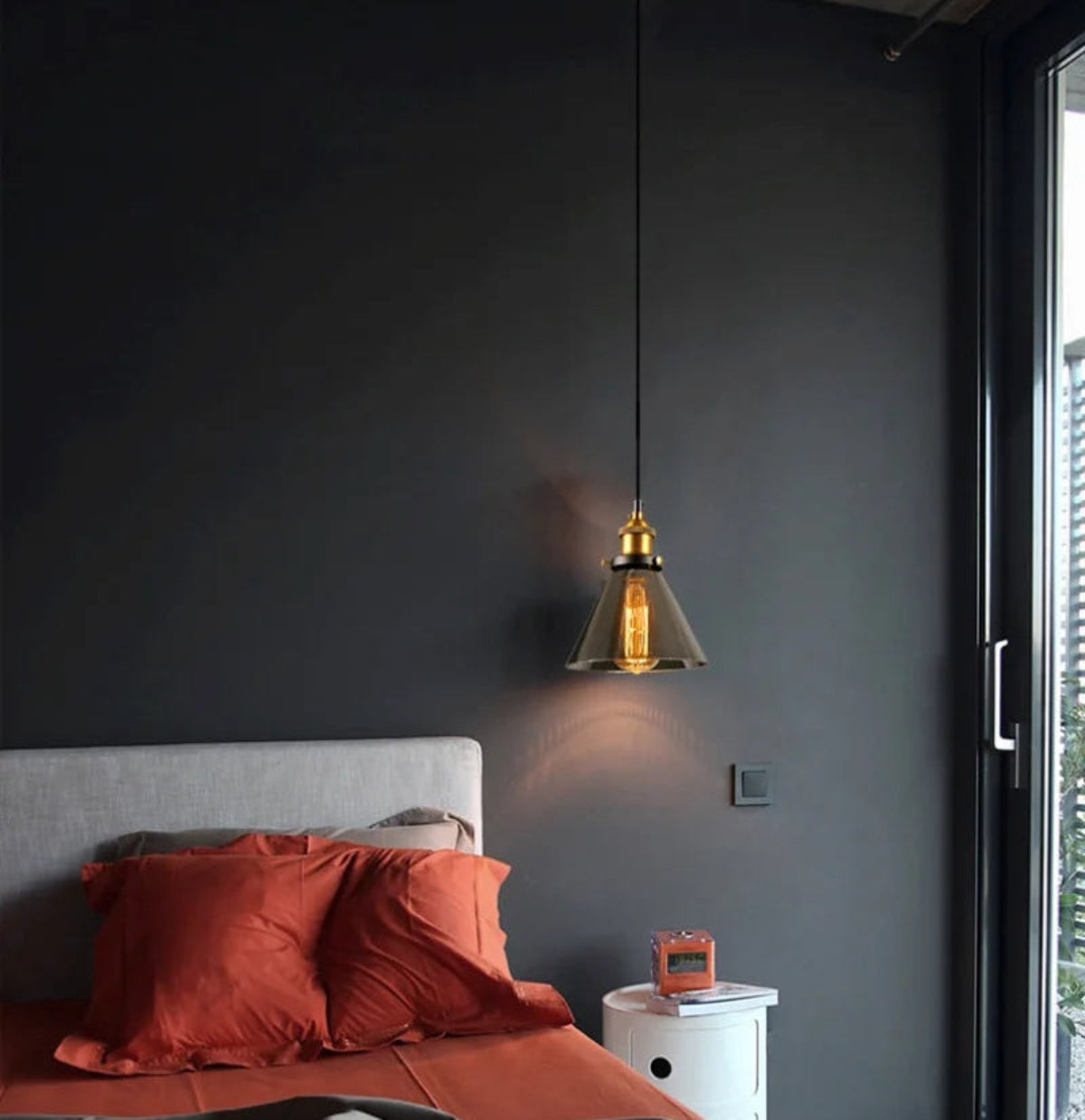 Smoke grey glass cone pendant light with brass fitting hung as a bedside light against a dark charcoal wall above a bed with terracotta linen bedding and a white nightstand — LivDecor
