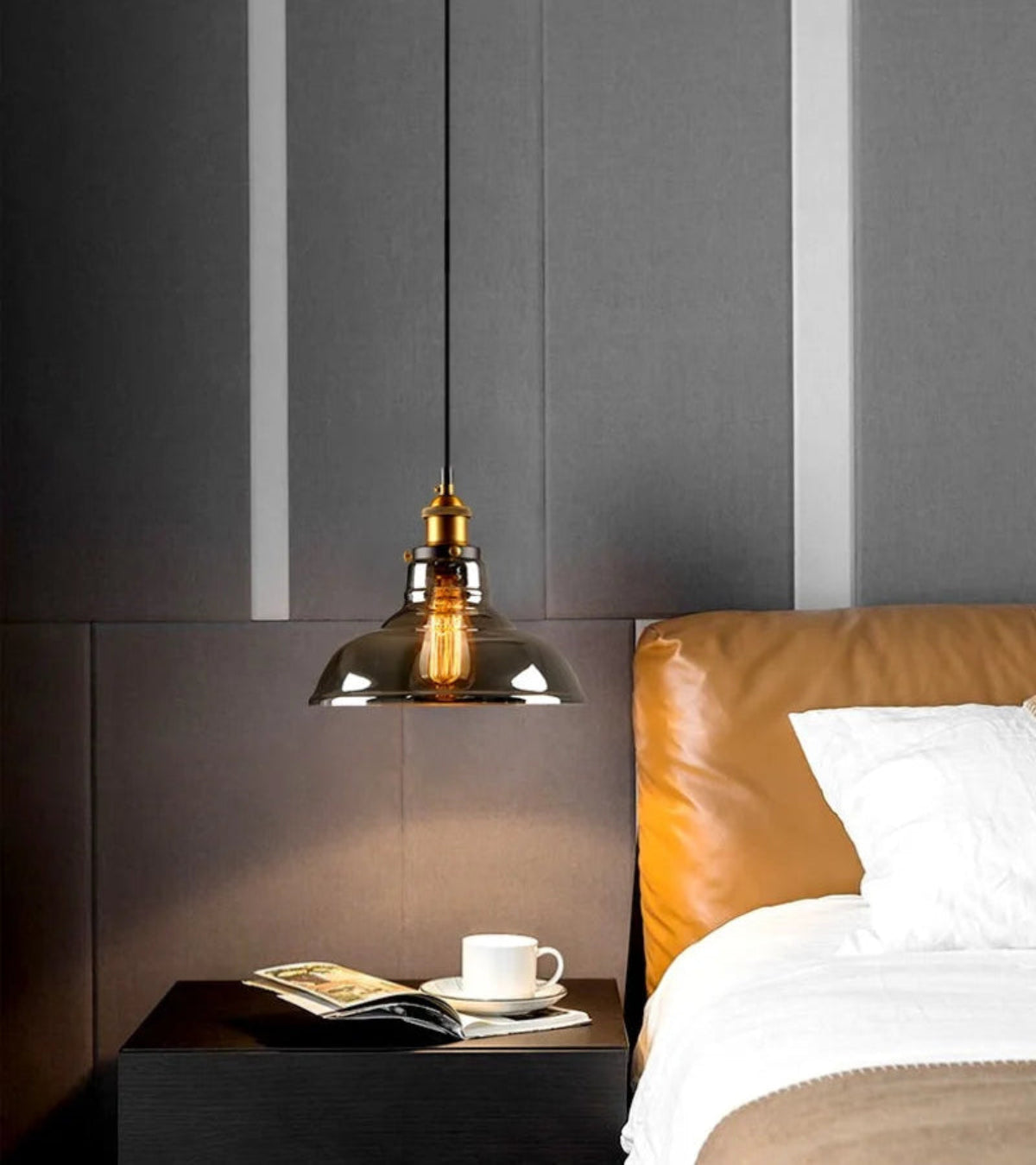 Smoke grey glass dome pendant light with brass fitting hung as a bedside light above a dark nightstand with an open magazine and white coffee cup, beside a tan leather headboard and white bedding — LivDecor