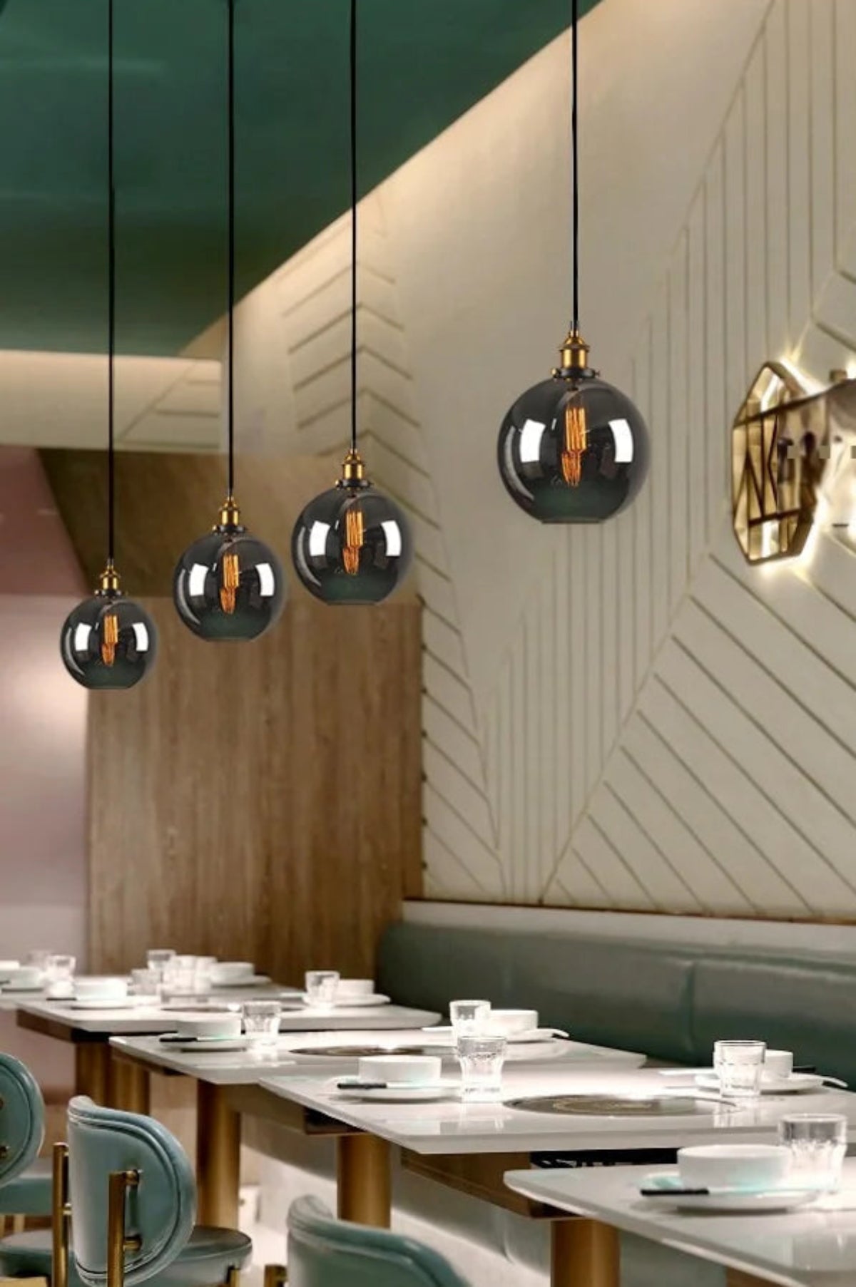 Four smoke grey glass globe pendant lights with brass fittings hung at varying heights above a long set dining table with green velvet banquette seating and geometric white paneled walls in a restaurant setting — LivDecor