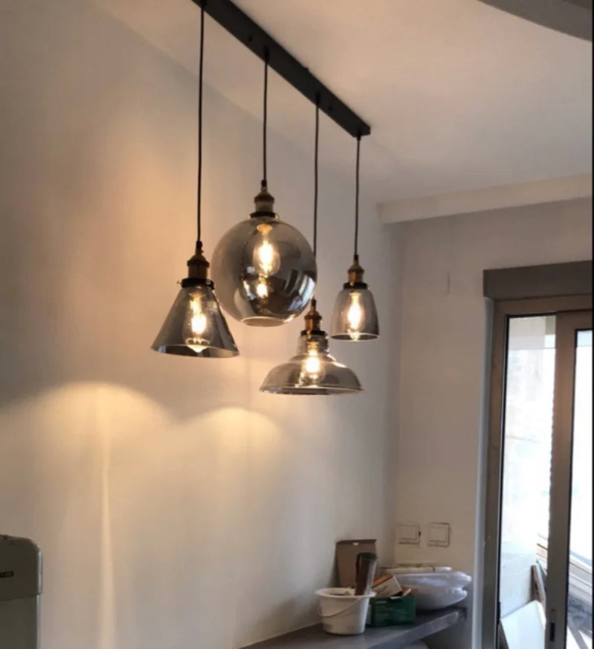 Five smoke grey glass pendant lights in globe, cone, dome and bell shapes hung at staggered heights from a black long bar ceiling plate above a kitchen counter in a real home installation — LivDecor