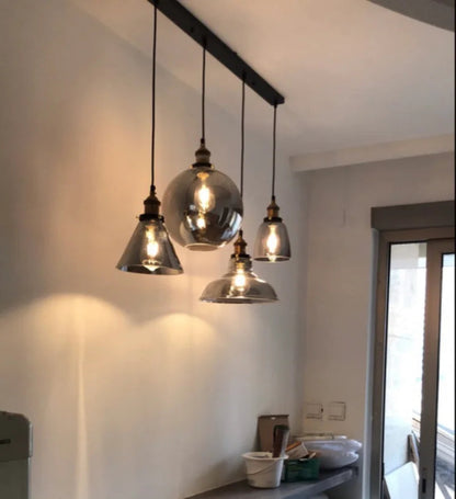 Five smoke grey glass pendant lights in globe, cone, dome and bell shapes hung at staggered heights from a black long bar ceiling plate above a kitchen counter in a real home installation — LivDecor