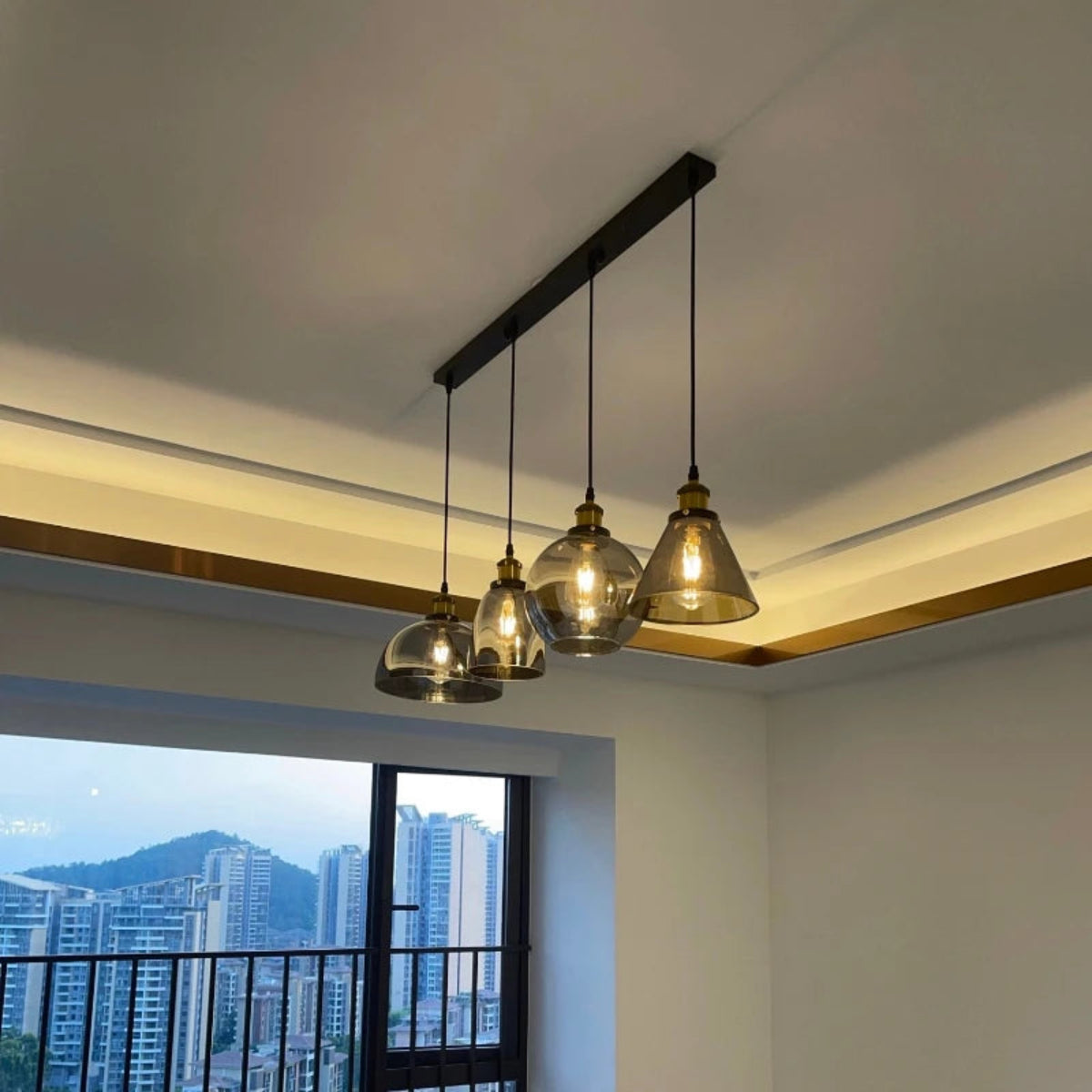 Three smoke grey glass pendant lights in dome and cone shapes on a black linear bar mount installed in a modern apartment with city skyline view — LivDecor