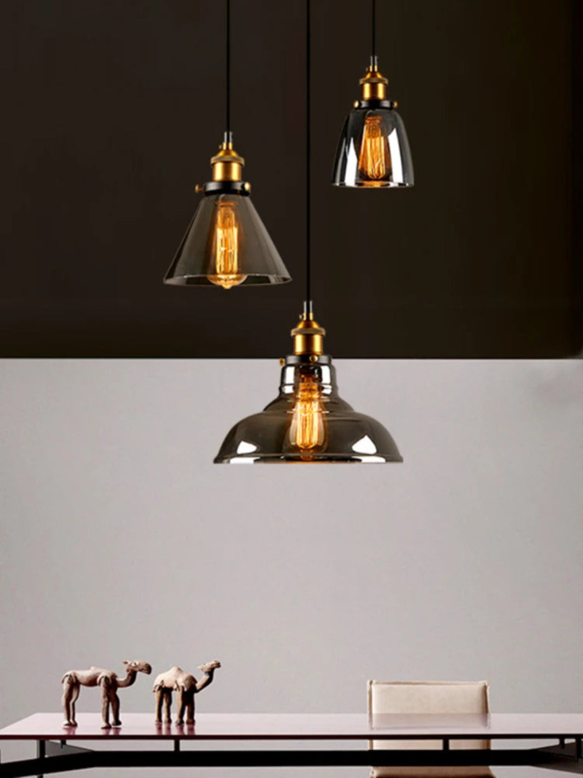 Three smoke grey glass pendant lights in cone, bell and dome shapes with brass fittings and glowing Edison filament bulbs against a dramatic dark and light split background above a minimalist dining table — LivDecor